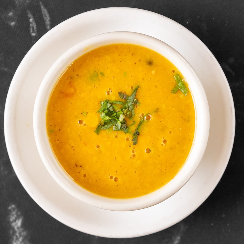 A white bowl of vibrant orange Indian lentil soup, garnished with fresh green herbs, on a dark surface.