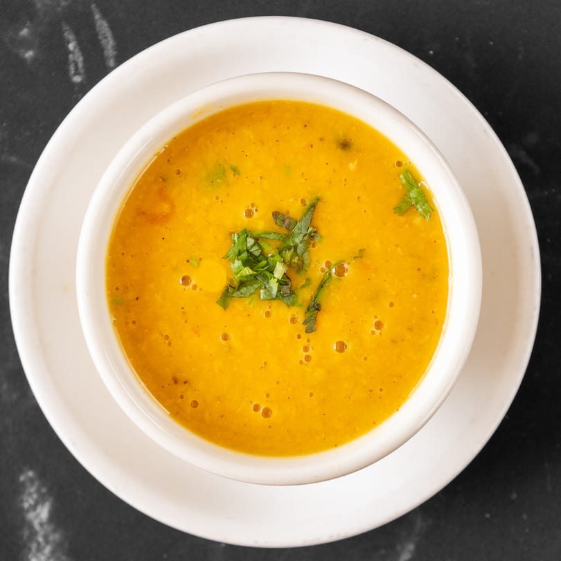 A white bowl of vibrant orange Indian lentil soup, garnished with fresh green herbs, on a dark surface.