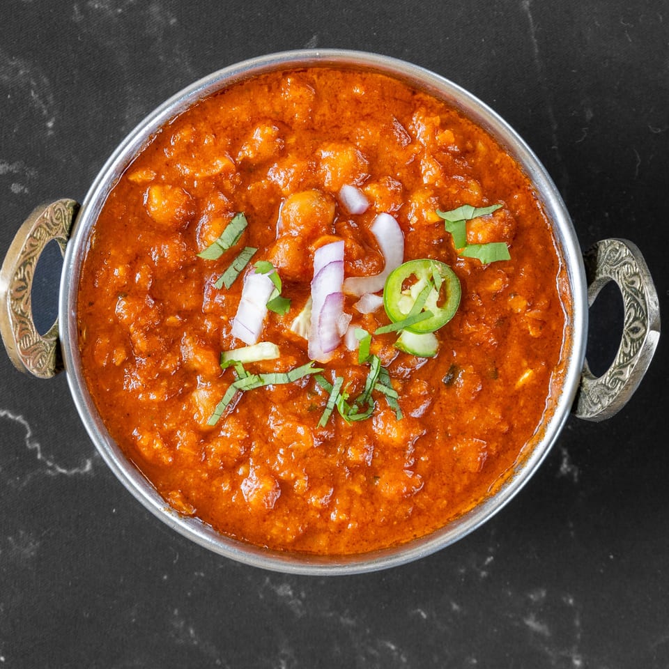 Overhead view of a metal bowl filled with Swad Indian Restaurant's chana masala, garnished with onions and chilies.