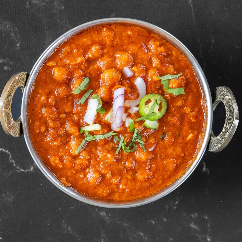 Overhead view of a metal bowl filled with Swad Indian Restaurant's chana masala, garnished with onions and chilies.