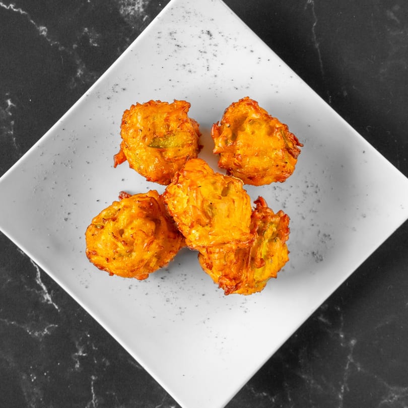 Five golden-brown Indian pakoras, or vegetable fritters, arranged on a white square plate.