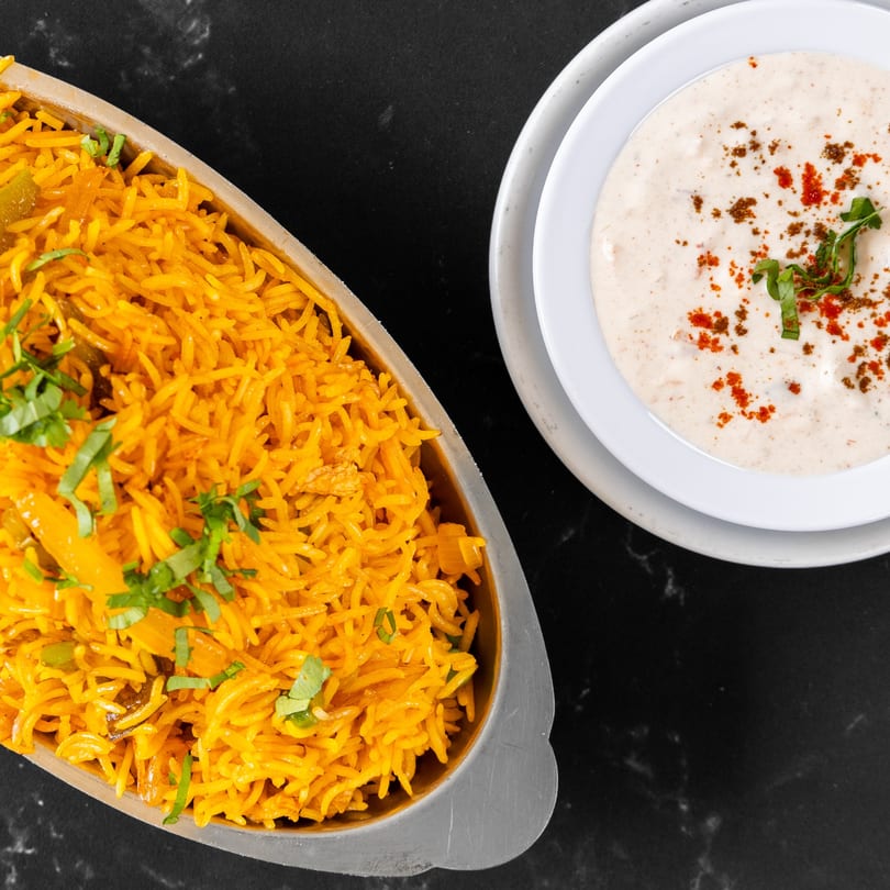 Overhead view of yellow Indian rice in a metal dish next to a white bowl of raita.
